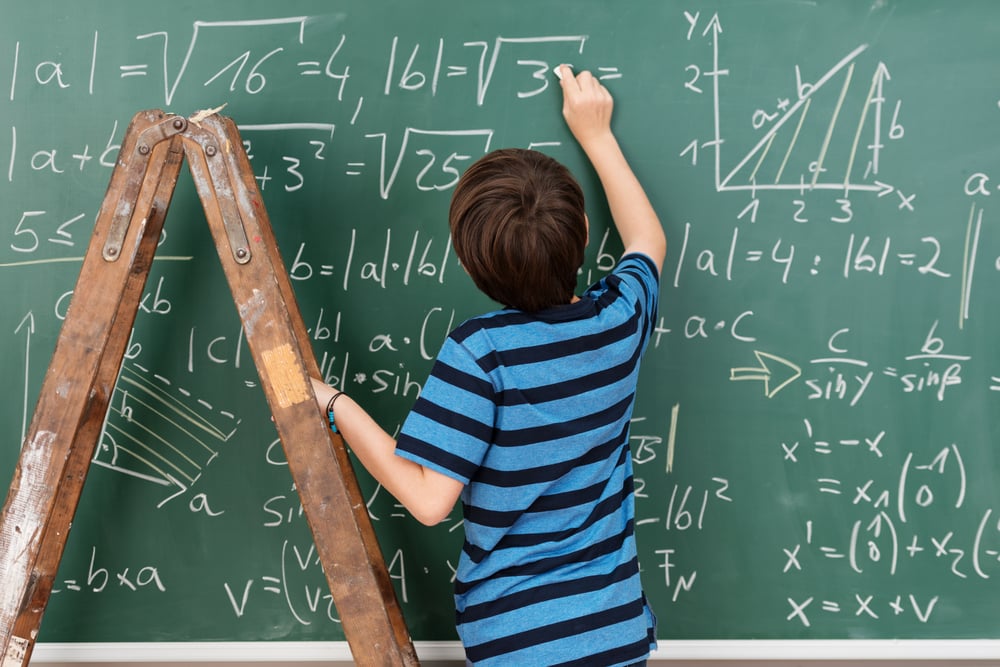 Highly intelligent little boy in the classroom standing on a stepladder to reach a complex mathematical problem on the blackboard that he is busy solving