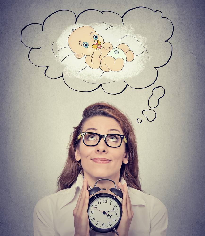 Portrait happy woman dreaming a baby looking up holding alarm clock isolated on gray wall background. Positive face expression emotion